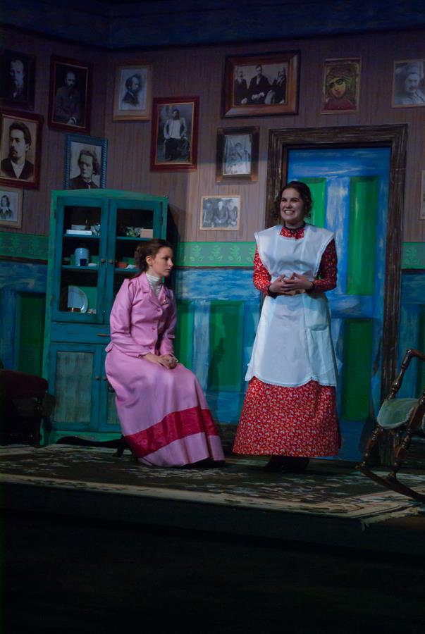 Two women in period costumes perform in a vintage living room set. One is standing in a red dress and apron, smiling, and the other sits in a pink dress, listening. The room has green walls and framed photos.