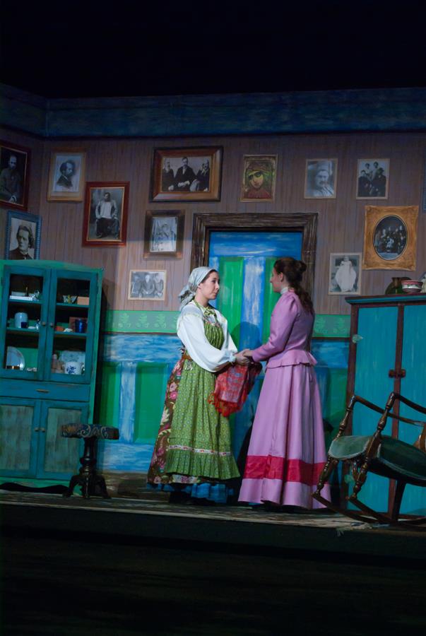 Two actors stand on a stage set as a vintage room. One wears a green floral dress and headscarf, the other a pink dress. They hold hands, surrounded by a rustic decor with picture frames and furniture, creating an old-fashioned atmosphere.