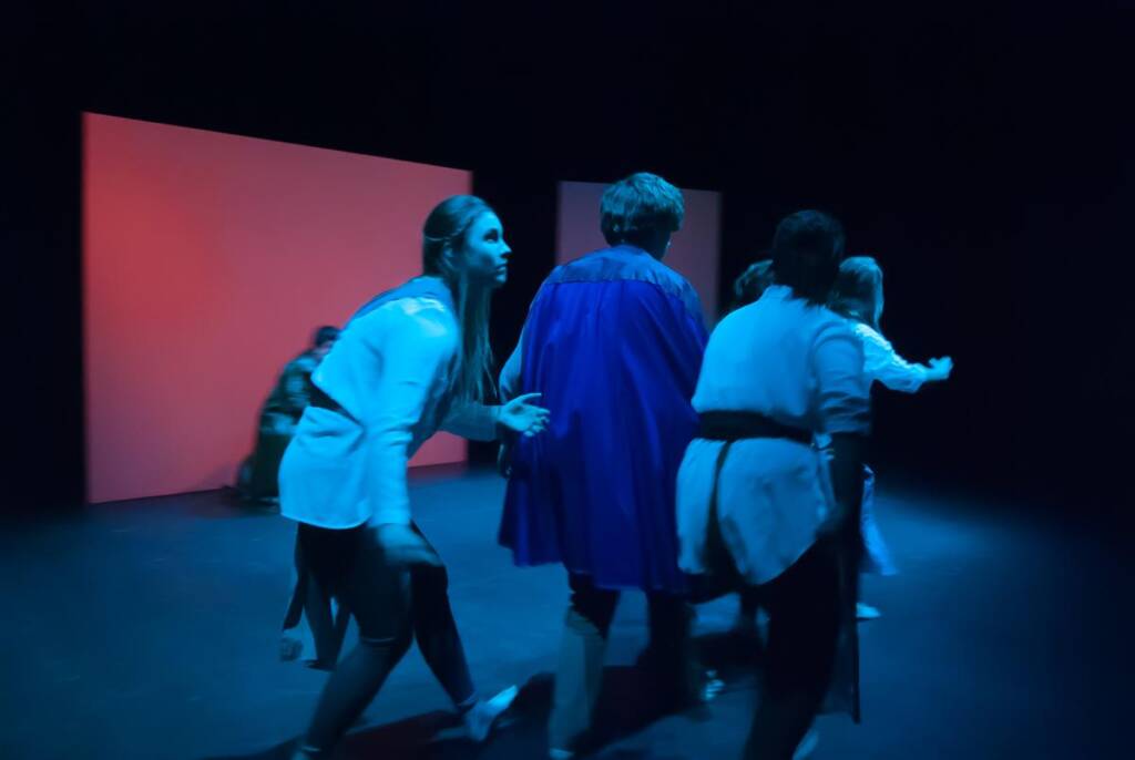 A group of actors performs on a dimly lit stage, dressed in medieval-style costumes. The background features a large, flat panel with red lighting, creating a dramatic atmosphere.