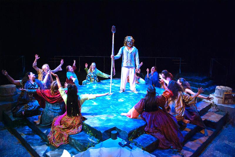 A theatrical performance with a central figure holding a staff on a blue-lit stage. Surrounding them are a group of people in colorful costumes, reaching out and directing their gaze towards the figure. The scene conveys a dramatic, mystical atmosphere.