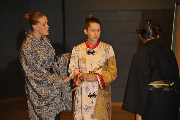 Three people in traditional costumes performing a play on stage. Two are wearing patterned robes with shiny details and buns, while the third has their back to the camera, dressed in a dark outfit. The setting appears to be indoors with a dimly lit background.