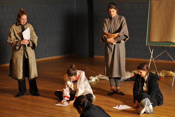 Four people in a room with wooden floors, three sitting and one standing. They appear focused and engaged in writing or discussion. The atmosphere seems to be related to a workshop or rehearsal.