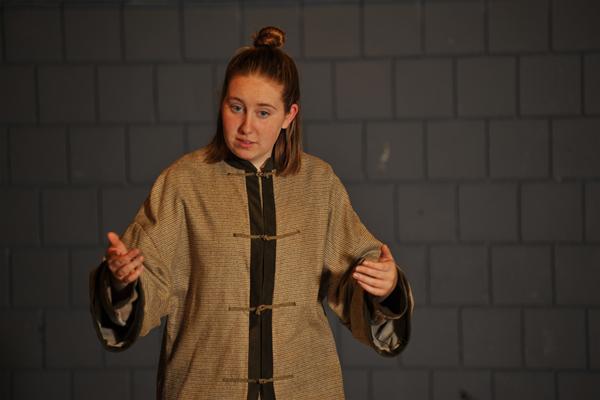 A person with medium-length hair is gesturing with both hands while wearing a long, buttoned coat. The background consists of a wall with rectangular, grayish tiles. The lighting is dim, focusing on the individual.