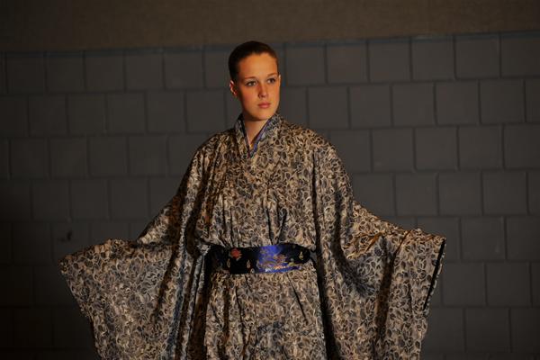 A person stands against a tiled wall wearing an ornate, patterned kimono with wide sleeves. The kimono features intricate designs, and the person holds their arms out to the sides, showcasing the garment's detail.