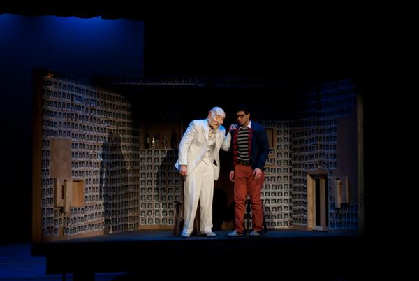 Two actors stand on a dimly lit stage with a backdrop resembling a room filled with shelves. One wears a white suit and glasses, gesturing while leaning forward. The other wears a striped shirt with a blazer and red pants, listening attentively.