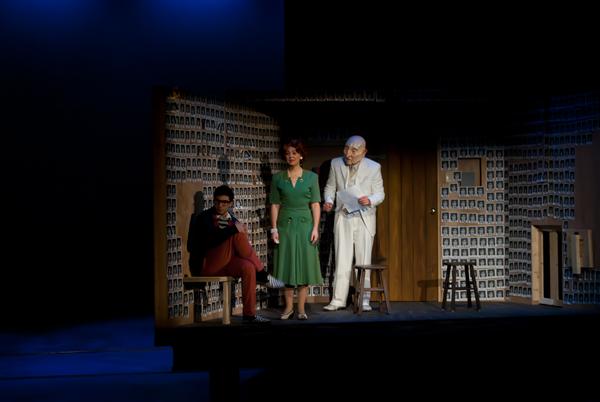 Three actors perform on a dimly lit stage in a room lined with photos. One sits on a stool reading, while the other two stand talking. The woman wears a green dress, and the older man is in a white suit. The backdrop creates a cozy, vintage ambiance.