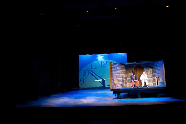 A stage with a large clock backdrop. Two actors sit on a small, wooden set piece resembling a living room. The lighting creates a blue ambiance, focusing on the actors, one in dark clothing and the other in white.