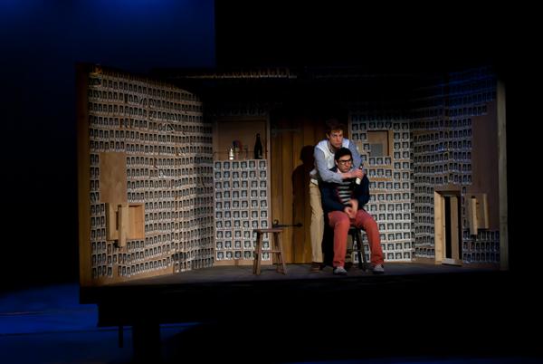 Two actors perform on a dimly lit stage, with one seated and the other standing behind. The set is a room lined with numerous small photographs. The seated actor wears glasses and red pants, while the other wears a blue shirt and vest.