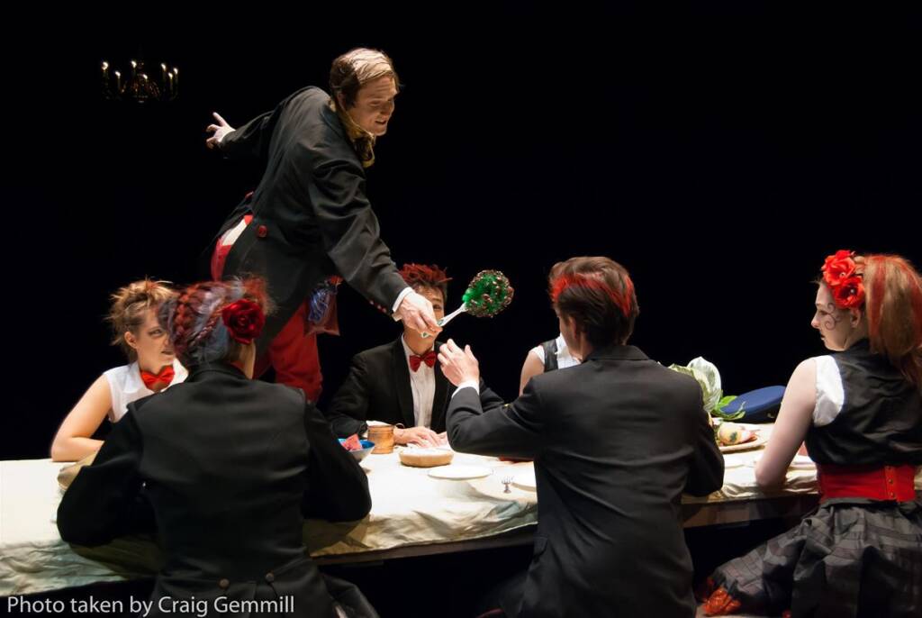 A theatrical scene with six actors in formal attire seated around a table. A man, standing on the table, presents a decorated skull to another actor. The setting is dramatic with dim lighting and focused spotlights.