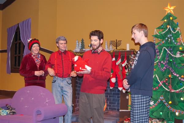Four people stand in a living room decorated for the holidays. They are near a Christmas tree and a fireplace with stockings. The group appears cheerful, with one person holding a holiday gift bag.