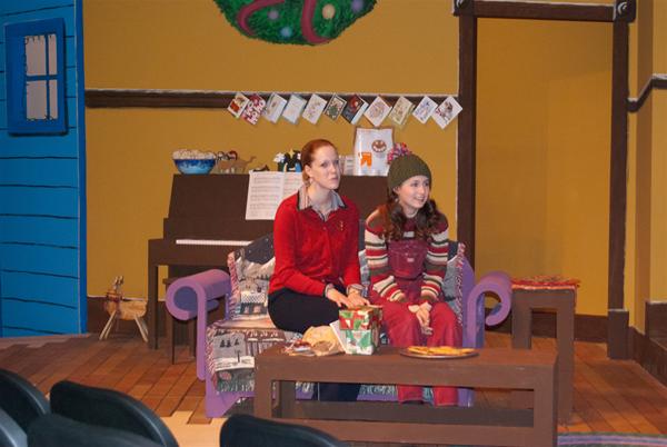 Two people sit on a purple couch in a festive living room. A piano with sheet music and holiday cards is in the background. The room is decorated, including a wreath on the wall. They seem to be on stage in a theater setting.
