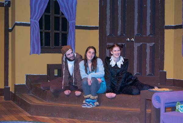 Three people in costumes sit on a stage set resembling a room with purple curtains and a wooden door. The scene is brightly lit, and they appear to be in mid-conversation, conveying a sense of whimsical storytelling.