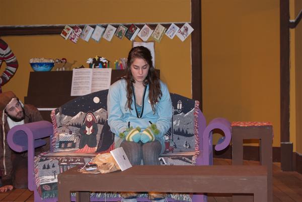 A woman sits pensively on a colorful couch with winter-themed patterns. Behind her is a wall decorated with holiday cards and a piano. A man peeks from behind the couch on the left. There is a small table with papers in front of her.