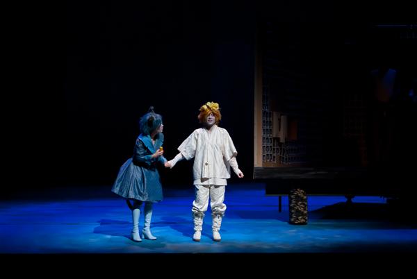 Two performers on stage are dressed in colorful, whimsical costumes with dramatic lighting. One wears a blue dress and the other a white outfit with orange accents. They appear to be engaged in a theatrical scene with dim lighting in the background.