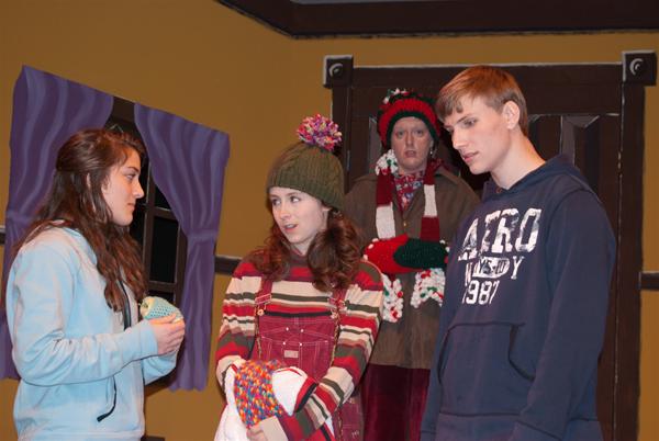 Four people in a room with yellow walls. One is wearing a light blue hoodie, another in overalls and a knit hat, a third in a winter coat with a pompom hat, and the fourth in a navy hoodie. There's a window with purple curtains.