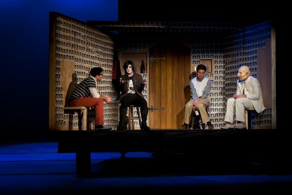 Four actors perform a skit on a stage with a backdrop resembling a room adorned with identical pictures. One actor in a dark outfit gestures, while the others sit or stand nearby in casual and formal attire, with focused expressions.