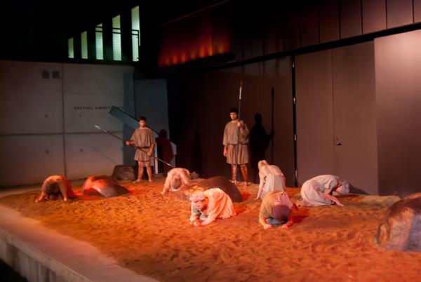 Dancers perform on a sand-covered stage, wearing beige costumes. Some kneel or sit in varied poses. The scene is dimly lit with orange hues, creating a dramatic atmosphere. Large rocks are scattered across the stage.