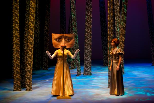 A performer on stage wears a costume with a large expressive face mask that covers their torso, standing amidst vertical scenic elements resembling tree trunks. Another person in a dress stands nearby, observing them. The lighting casts a colorful ambiance.