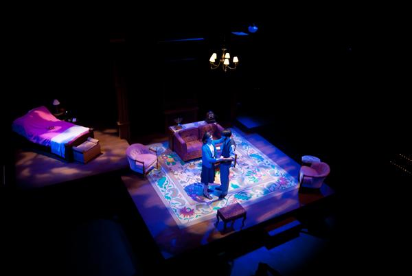 A dimly lit stage with a couple dancing in the center. The set resembles a cozy living room with a colorful rug, a couch, armchairs, and a bed in the background. Warm lighting highlights the intimate scene.