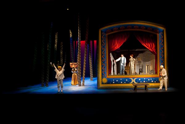 A dimly lit stage with actors in elaborate costumes. One actor is in a colorful, decorated frame resembling a puppet theater. Another actor stands in the foreground with arms raised. Tall, thin set pieces add to the dramatic effect.