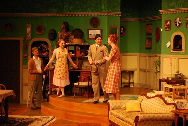 Four actors on a stage set of a living room during a play. One man in a suit and hat is shaking hands with a smiling woman in a plaid dress. Two other people stand nearby, one woman in an apron and one older man with a cane.