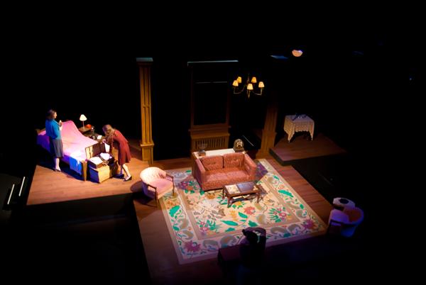 An overhead view of a theatrical stage set depicting a living room. It includes a sofa, chairs, a coffee table, and a decorative rug. Two people are interacting near a bed on the left side. The scene is dimly lit, creating a dramatic ambiance.