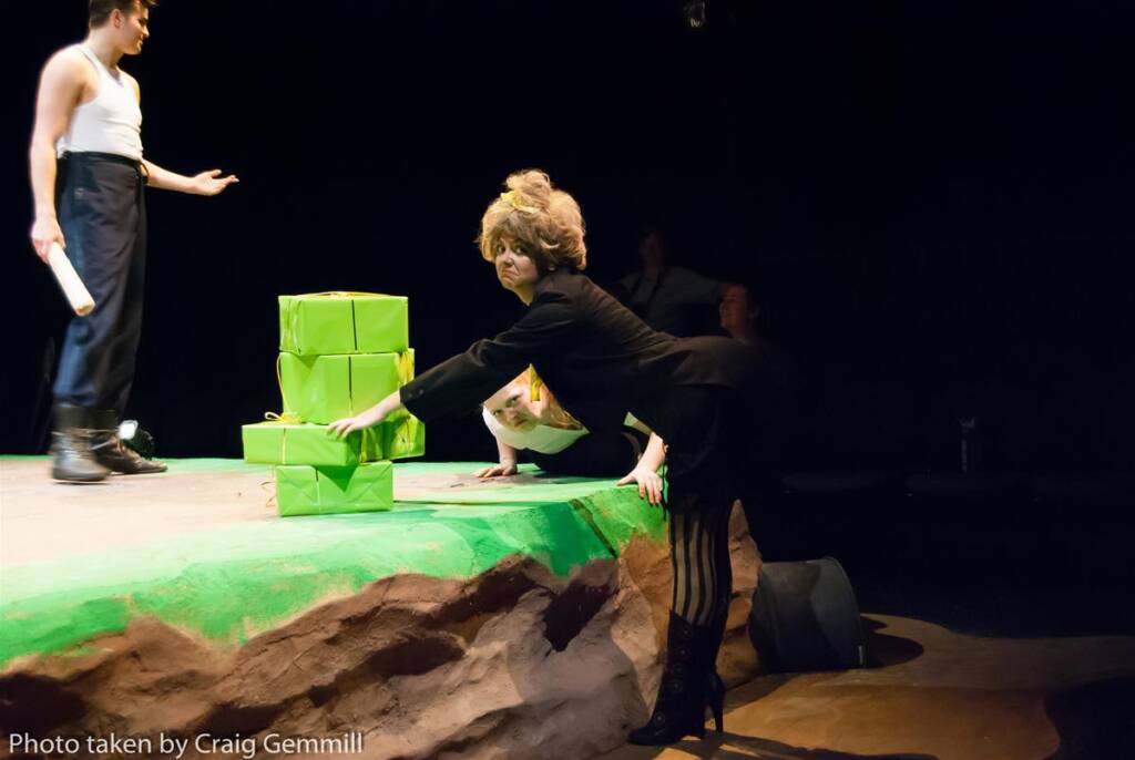 A person in a suit and boots is playfully reaching for a pile of wrapped green gifts on a stage. Another person in a tank top stands nearby, partially out of view. The background is dark, with a rocky surface in the foreground.