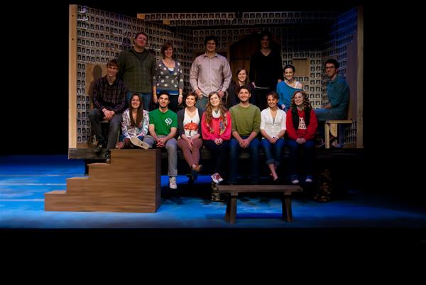 A group of people, some sitting and others standing, pose on a stage set with a patterned backdrop. The lighting is bright, focusing on the group. There are stairs and a small bench on the set. Everyone is casually dressed.