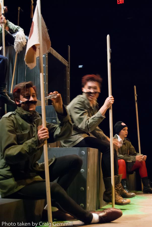 Performers dressed in military-style costumes sit on tiered metal structures in a theater setting. They hold long sticks and some have fake mustaches. Dim lighting casts shadows. Red exit sign glows in the background.