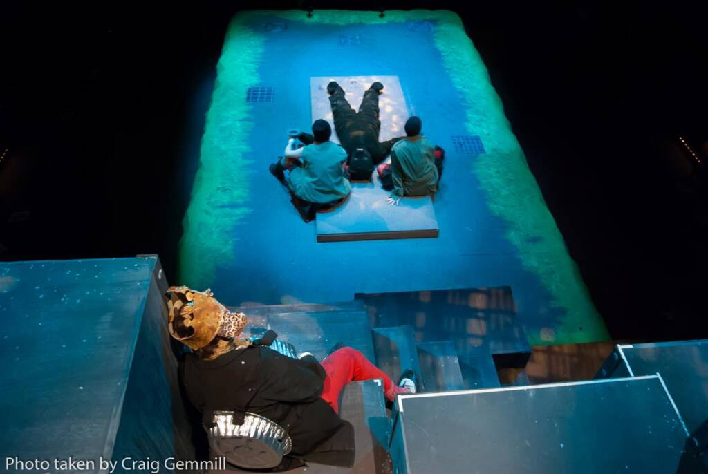 A theatrical performance scene viewed from above, featuring four actors on a stage. One actor lies on the floor, while three others kneel near him. The set is illuminated by blue and green lighting. A person in costume sits apart, observing.