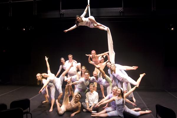 A group of performers in soft purple and white attire strike dynamic poses on a stage. One person is suspended in the air using a long fabric, while others form a pyramid-like structure below. They are all smiling and exude a sense of joy and unity.
