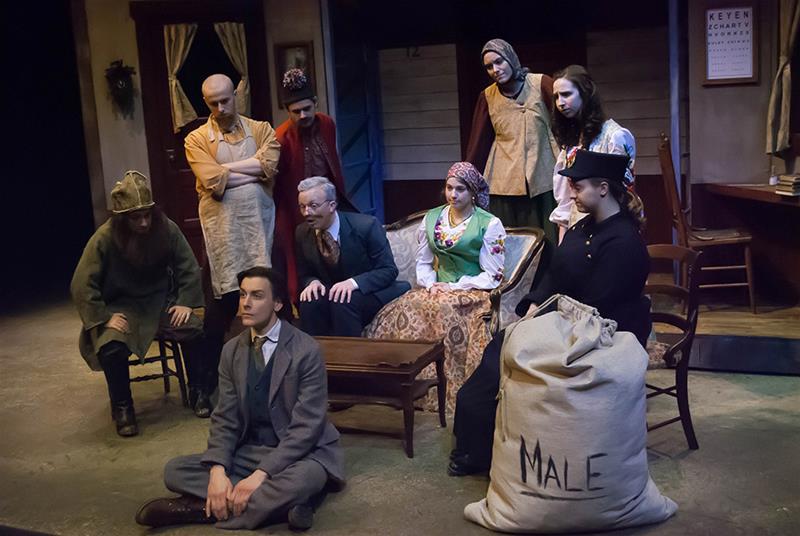 A group of actors in period costumes perform a dramatic scene in a theater setting. They are gathered around a central male figure in a suit, with some sitting and others standing. A large sack marked "MALE" is in the foreground.