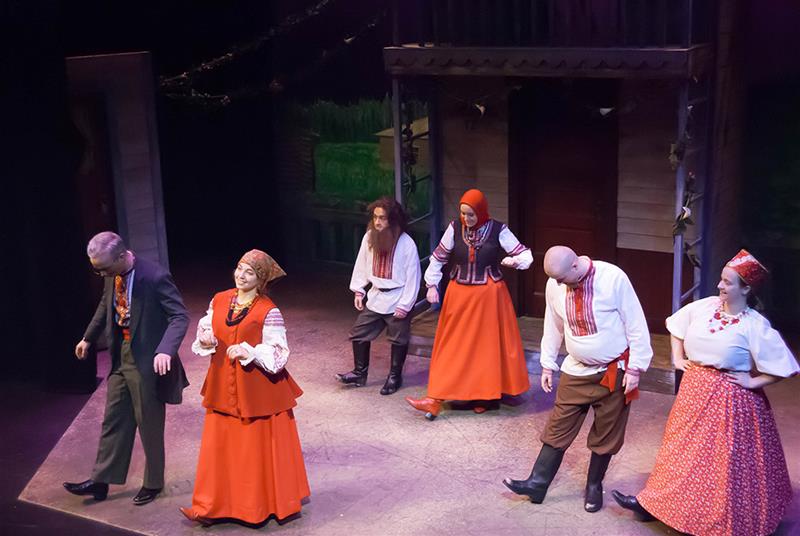 A group of performers on stage in traditional folk costumes, including long skirts and headscarves. They are mid-performance, with a set featuring a wooden structure in the background. The scene is warmly lit.