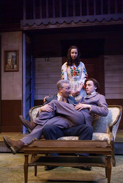 Three actors perform on stage. Two sit closely on an ornate sofa, one reclining against the other, while the third stands behind them with a neutral expression. The setting includes a painting and wooden backdrop, suggesting a cozy, indoor scene.