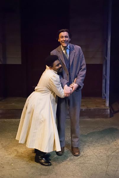 Two people on stage embrace and smile. One is wearing a light-colored coat and leaning against the other, who is in a suit and tie. The background is dimly lit, suggesting a theatrical setting.