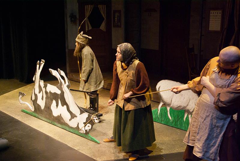 Theater actors in rustic costumes perform a play. One actor holds a cutout of a cow, while another guides a cutout of a sheep. The setting suggests a countryside scene with dim lighting.