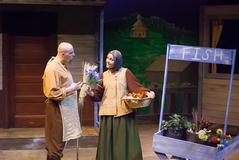 A man and woman in period costumes engage in conversation onstage. The man holds flowers, and the woman, wearing a headscarf, carries a basket. Beside them is a "FISH" stand with potted plants. A rustic, village-like set decorates the background.