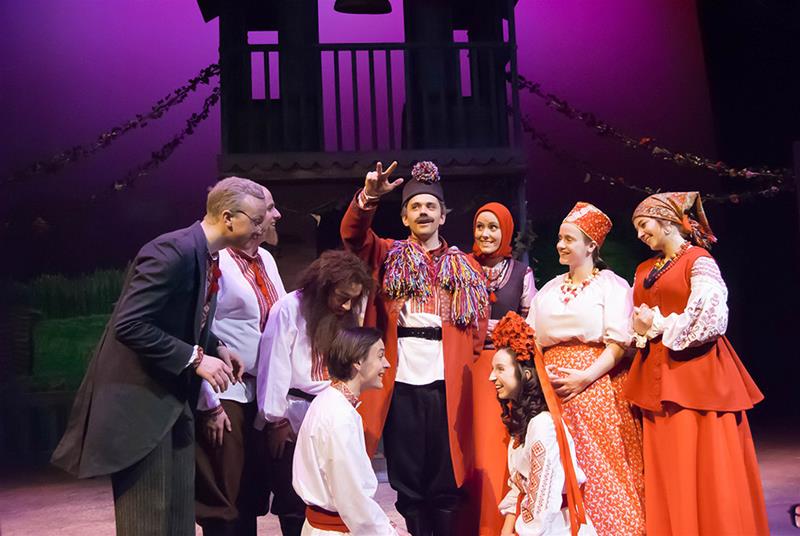 A group of people in colorful, traditional Eastern European costumes are joyfully gathering on stage. One person in the center is gesturing upwards, while others around them watch and smile. The backdrop shows a house and festive decorations.