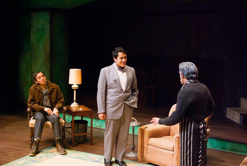 Three actors perform on a stage. One actor sits relaxed in a chair, while another stands in a gray suit, engaged in conversation with a third actor wearing a dark sweater and striped apron. A lamp on a side table lights the scene.
