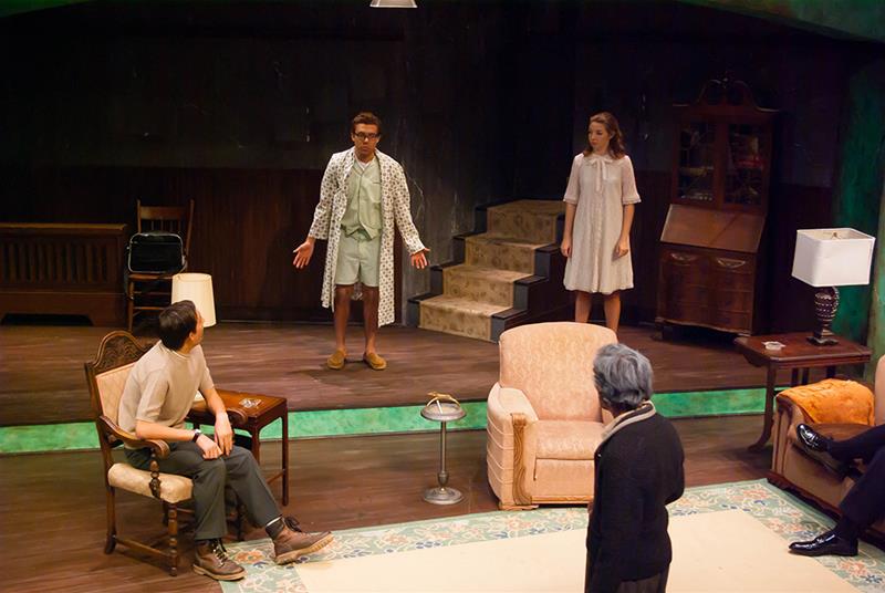 A group of actors perform a scene in a living room set on stage. One stands in the center wearing a bathrobe, while two others in casual clothing and one seated elder face them. The scene conveys emotion and tension.