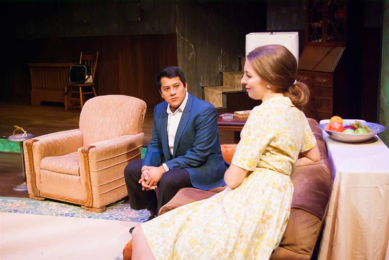 A man in a blue suit sits on a couch next to a woman in a yellow dress. They appear to be engaged in a conversation. The setting is a cozy living room with a rug, armchair, and small table with fruit.