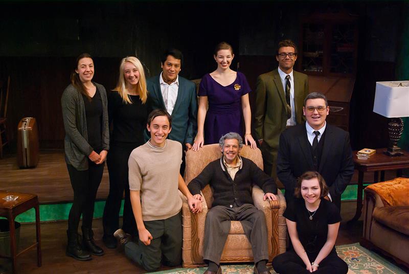 A group of ten people are posing for a photo in a cozy room. Four people are standing, three are kneeling, and one is seated in an armchair, holding hands with two kneeling individuals. The room has a lamp and various furniture.