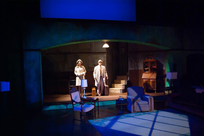 A dimly lit stage set resembling a cozy living room with vintage furniture, including two armchairs and a lamp. Two actors stand under a spotlight near the center, dressed in period clothing, conveying a dramatic scene.