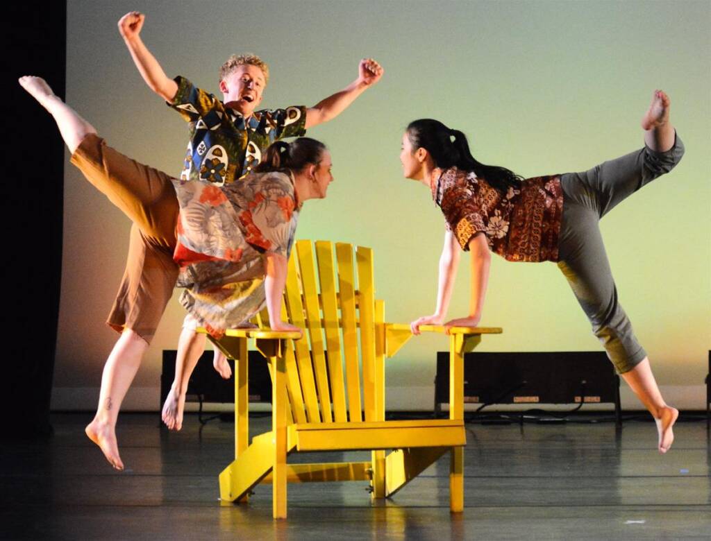 Three dancers perform energetically on and around a yellow wooden chair. They wear colorful shirts and pants, with two supporting themselves on the chair's arms and one standing energetically behind, arms raised. The setting is a stage with soft lighting.