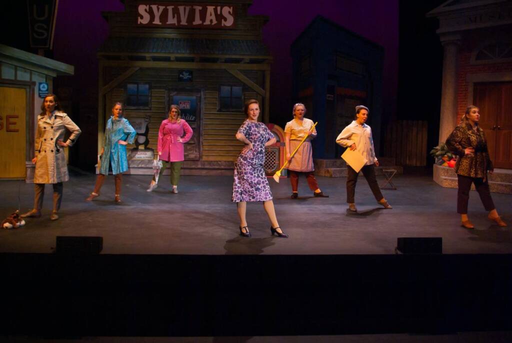 Seven performers on stage in colorful costumes strike dramatic poses in front of a set resembling a small town with a wooden building labeled "Sylvia's." The lighting is warm, highlighting the lively scene.