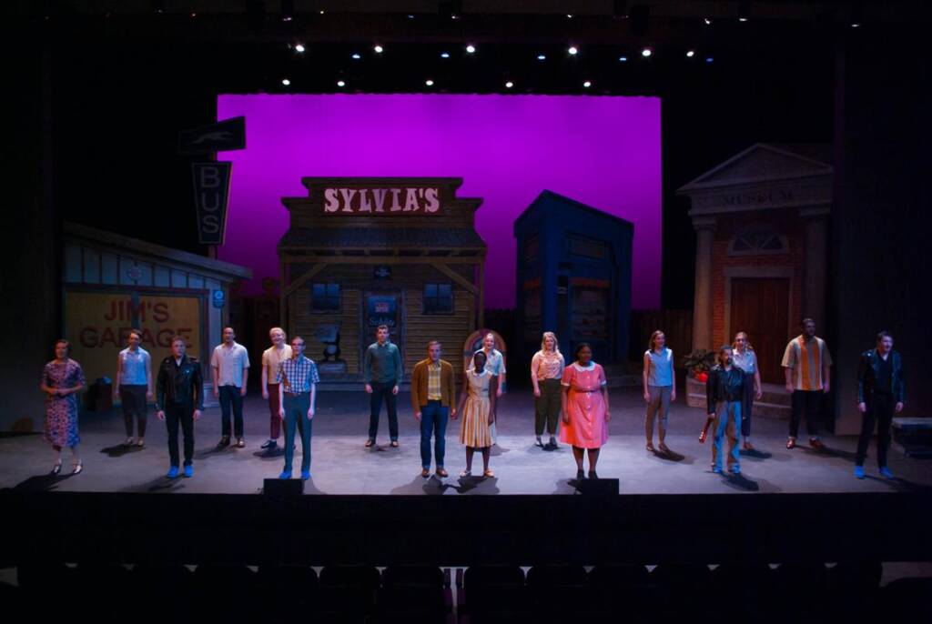 A group of actors stand on stage in front of a set featuring buildings labeled "Sylvia's" and "Jim's Garage." The backdrop is lit with pink and purple lighting. The cast is spread across the stage, each in various costumes.