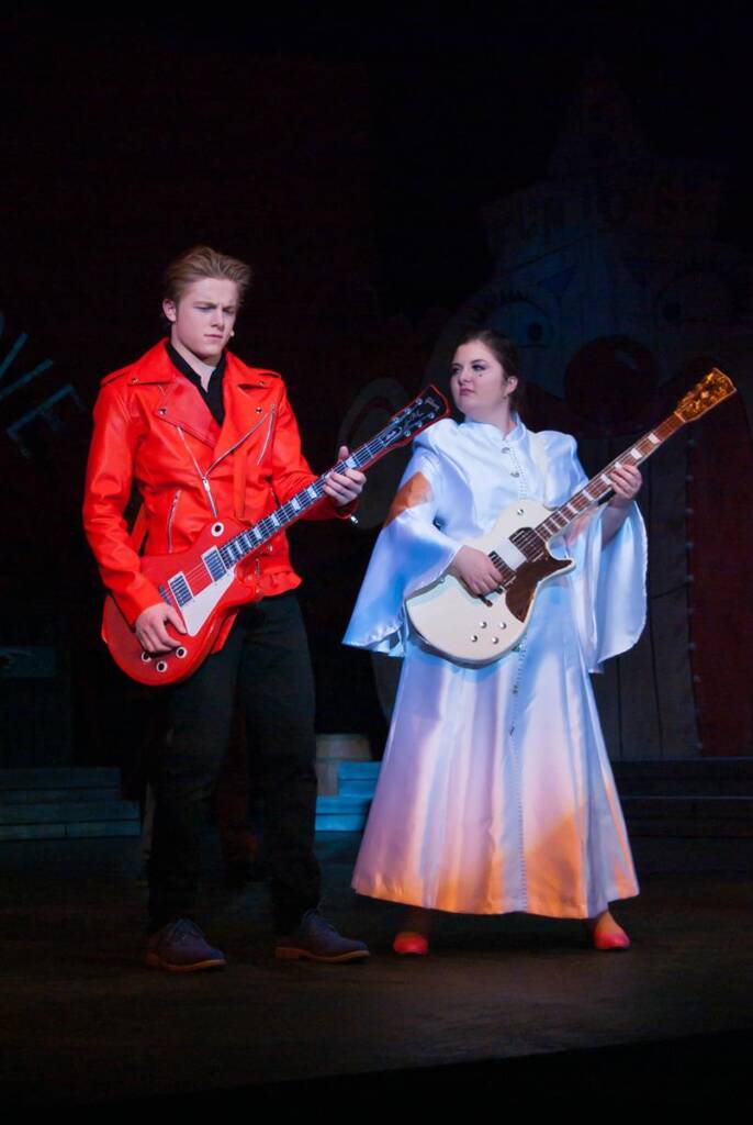 A person in a bright red jacket plays an electric guitar next to a person in a white dress also playing an electric guitar. They are performing on stage under dim lighting.