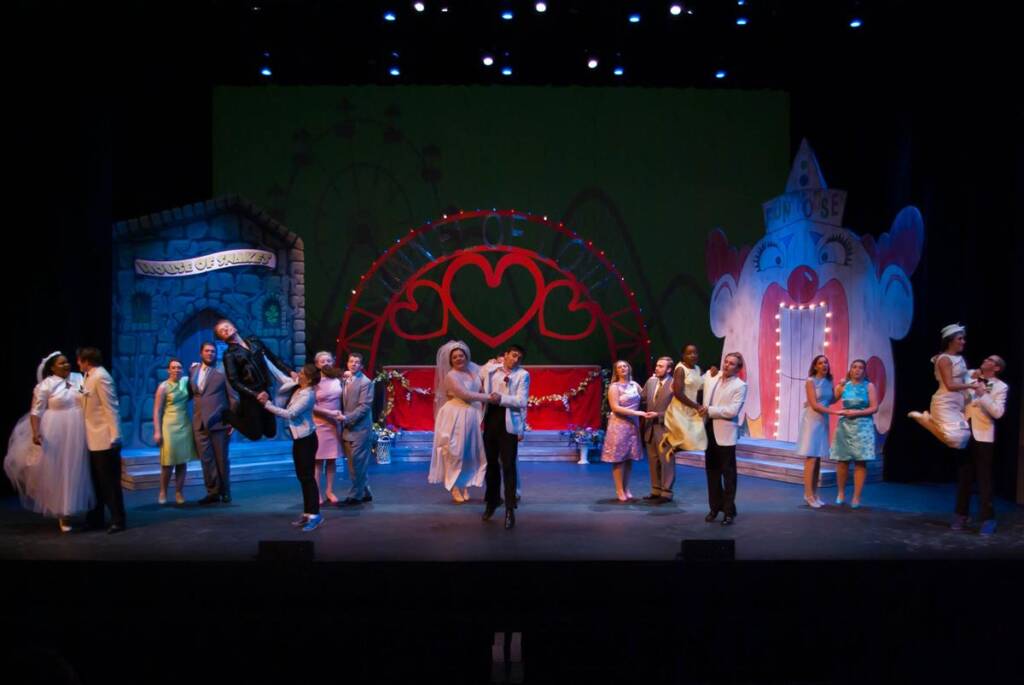 A vibrant stage performance depicting a lively scene with multiple couples dancing. The backdrop features a colorful, whimsical carnival theme with bright lights and heart motifs. Performers are in various costumes, creating a festive atmosphere.