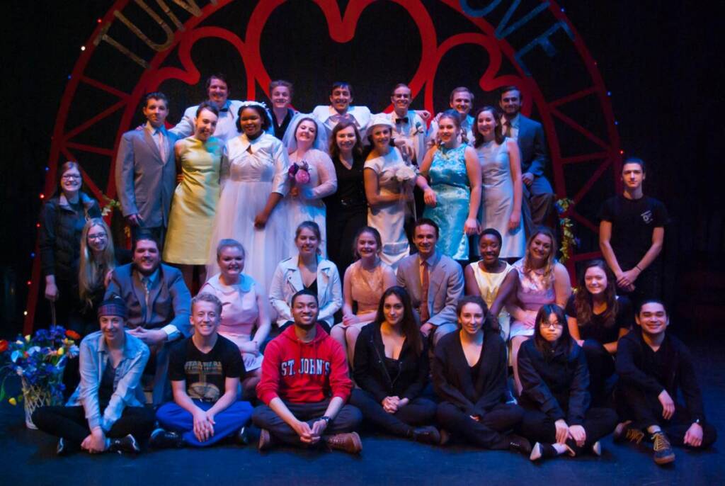 A large group of people poses on a stage, with some standing and others sitting or kneeling. They are dressed in a variety of outfits, suggesting a theater production. A decorative arch with a heart design is in the background.