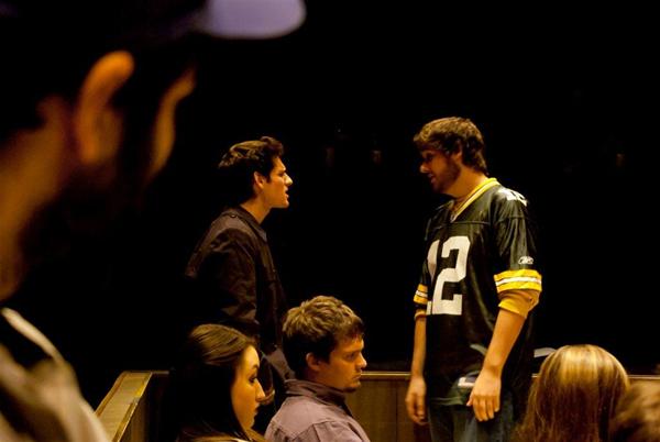 A group of people is inside a dimly lit room. Two men are standing and facing each other, with one wearing a number 12 sports jersey. Others are seated, attentively watching their interaction. A person in a cap is in the foreground.